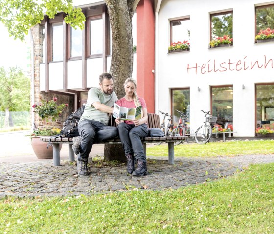 Two people are sitting on a bench in front of the Heilsteinhaus in Einruhr, reading a brochure. Bicycles are parked in front of the building, which is decorated with flowers., © -@Eifel Tourismus GmbH, AR-shapefruit-AG