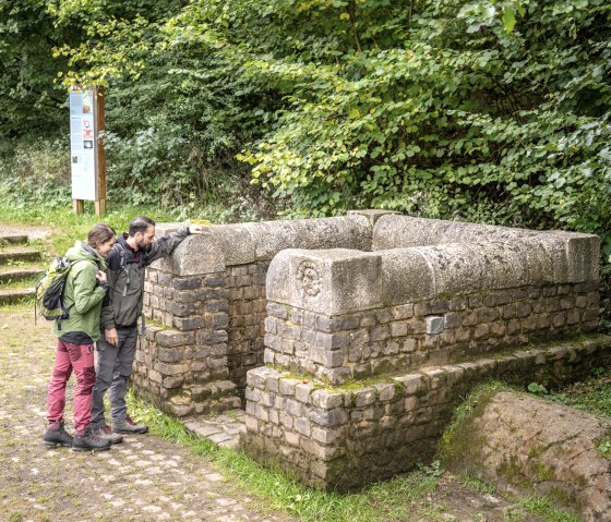 Zwei Personen betrachten einen alten Brunnen aus Stein in einem Waldgebiet. Im Hintergrund sind Bäume und ein Informationsschild zu sehen., © Eifel Tourismus GmbH, Dominik Ketz