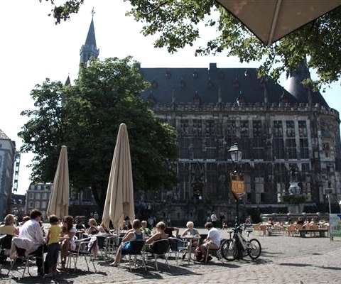 Menschen sitzen in einem Straßencafé vor dem historischen Rathaus in Aachen. Das Gebäude ist von Bäumen umgeben, der Himmel ist klar., © Archiv StädteRegion Aachen