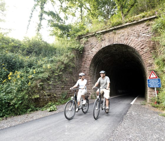 Zwei Radfahrer fahren aus einem bewachsenen Tunnel auf der Vennbahn bei Lommersweiler. Ein Warnschild steht am Rand., © vennbahn.eu