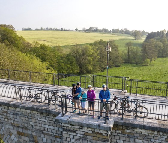 Gruppe von Radfahrern steht auf dem Rollefbachviadukt, umgeben von grüner Landschaft und Bäumen. Fahrräder lehnen am Geländer., © StädteRegion Aachen