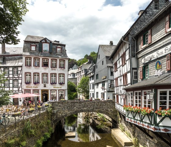 Fachwerkhäuser in Monschau mit einer Steinbrücke über einen Bach. Menschen sitzen draußen in einem Café. Blumen schmücken die Fenster., © Eifel-Tourismus GmbH, Dominik Ketz