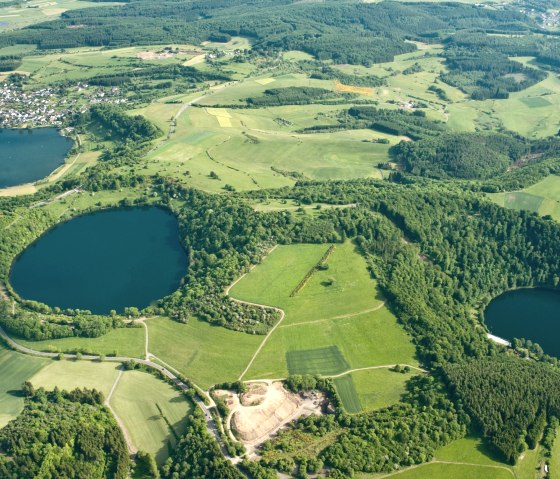Aerial view of the Dauner Maare in the Eifel. Three circular lakes are surrounded by green fields and forests. A small settlement is visible., © Gesundland Vulkaneifel, D. Ketz
